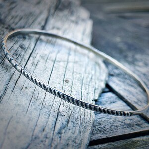 Könnte beinhalten: Ein silbernes Armreif-Armband mit einem strukturierten, seilartigen Design. Das Armband liegt auf einer verwitterten Holzoberfläche.