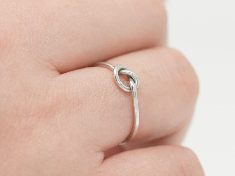 May include: A silver-colored ring with a knot design. The ring is thin and delicate, with the knot forming the focal point. The ring is worn on a finger.