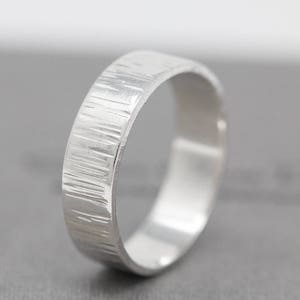 May include: A silver band ring with a textured, brushed finish. The ring is wide and has a simple, modern design.