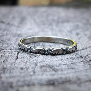 Può includere: Un anello d'argento con un motivo dettagliato di forme ripetute a forma di foglia. L'anello è posto su una superficie di legno grigio e invecchiato, che ne evidenzia il design complesso. La fascia dell'anello è sottile e delicata.