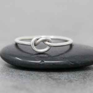 May include: A silver-colored ring with a knot design. The ring is set on a dark, smooth surface. The knot is the focal point, with the band appearing thin and delicate. The ring is a simple, elegant piece of jewelry.