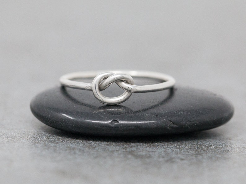 May include: A silver-colored ring with a knot design. The ring is set on a dark, smooth surface. The knot is the focal point, with the band appearing thin and delicate. The ring is a simple, elegant piece of jewelry.