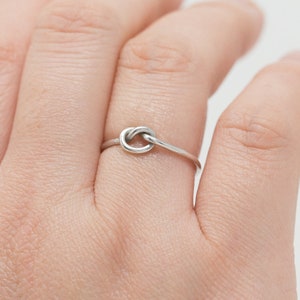 May include: A silver-colored ring featuring a knot design. The ring is thin and delicate, with the knot as the focal point. The ring is worn on a finger.
