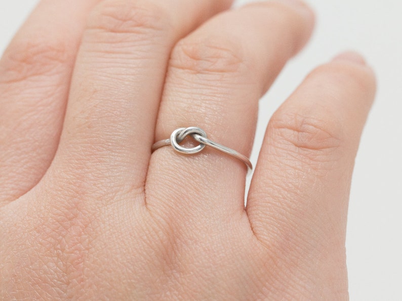 May include: A silver-colored ring featuring a knot design. The ring is thin and delicate, with the knot as the focal point. The ring is worn on a finger.