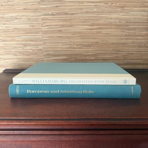 May include: Two hardcover books stacked on a dark wood surface. The top book has a light blue cover with a white spine and the title "Williamsburg Decorating With Style." The bottom book is teal with the title "European and American Dolls."