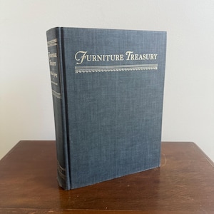 May include: A hardback book titled "Furniture Treasury" stands upright. The book has a textured, dark blue cover with the title in elegant script. The spine displays the title and author. The book is on a wooden surface.