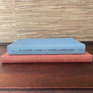 May include: Two vintage books stacked on a wooden surface. The top book is light blue with the title "Dictionary of Marks / Pottery and Porcelain." The bottom book is red with text. The background is a textured, light brown wall.