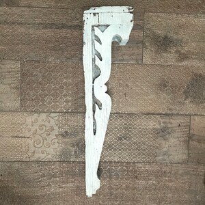 May include: A distressed, white-painted wooden corbel with decorative cutouts. The architectural salvage piece has a weathered appearance, showcasing its age and history. The corbel is designed to support a shelf or other structure.