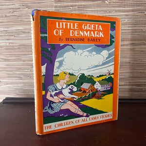 Peut inclure: Une couverture de livre vintage avec un fond orange et une illustration colorée d'une fille tenant un chien. Le titre est "Little Greta of Denmark" par Bernadine Bailey. Le livre fait partie de la série "The Children of All Lands Stories".