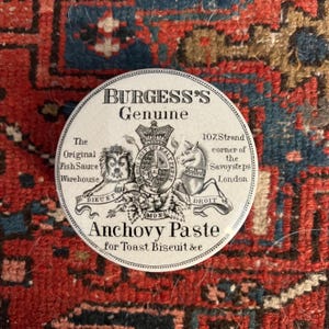 May include: A white ceramic jar lid with black lettering and a black and white illustration of a coat of arms. The text reads "Burgess's Genuine The Original Fish Sauce Warehouse 107, Strand corner of the Savoy Steps London Anchovy Paste for Toast Biscuit &c".