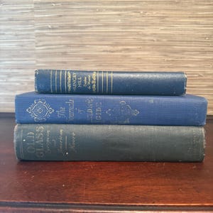 May include: Three antique books stacked on a wooden surface. The top book is dark blue with gold lettering and the title "Hungry Hill". The middle book is blue with the title "The Journals of Andre Gide". The bottom book is green with the title "Old Glass".
