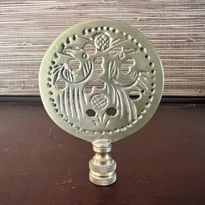 May include: A silver metal lamp finial with a round, decorative top featuring a cut-out design of stylized flowers and leaves.