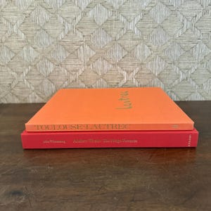 May include: Two hardcover books stacked on top of each other. The top book is bright orange with the text "TOULOUSE-LAUTREC" in gold on the spine and "lautrec" on the cover. The bottom book is red with gold text on the spine reading "Andrew Wyeth: The Helga Pictures".