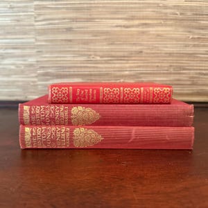May include: A stack of three antique books in varying shades of red. The top book is a smaller size with gold embossed detailing. The two larger books have gold lettering and decorative elements. The books are stacked on a dark wooden surface.