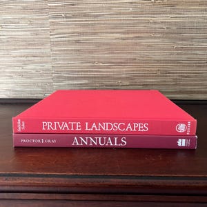 Peut inclure: Deux livres reliés en rouge, l'un sur l'autre. Le livre du haut est intitulé "PRIVATE LANDSCAPES" par Seibold & Sykes. Le livre du bas est intitulé "ANNUALS" par Proctor & Gray.