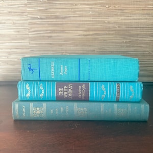 May include: Three vintage books stacked on a dark wooden surface. The books are teal and turquoise, with gold lettering and decorative elements. The top book reads "Ulysses" by James Joyce. The middle book is "The White Company" by Sir Arthur Conan Doyle.