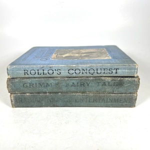May include: A stack of three vintage hardcover books with blue covers. The top book is titled "Rollo's Conquest", the middle book is titled "Grimm's Fairy Tales", and the bottom book is titled "Raggedy Ann's Entertainments".