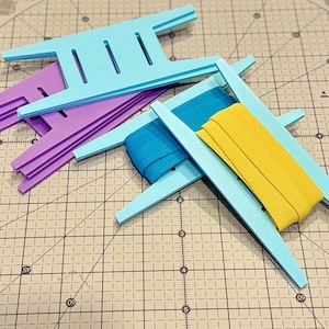 May include: A collection of craft tools, including light blue and purple foam ribbon holders, and spools of blue and yellow elastic ribbon. The tools are arranged on a grid cutting mat.