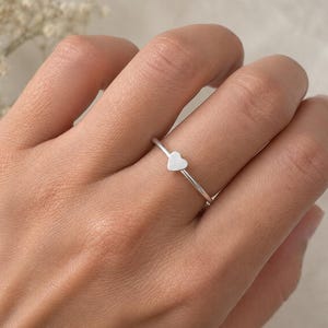 May include: A thin, silver-coloured ring featuring a small, flat heart-shaped charm centred on the band. The metal has a smooth, matte finish, and the band is slender, creating a simple, understated look on the finger without any additional details.