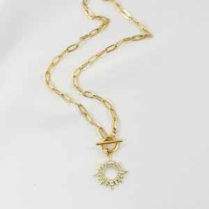 Peut inclure: Collier doré avec un pendentif en forme d'éclat de soleil. La chaîne est composée de maillons ovales allongés et d'un fermoir à bascule. Le pendentif est un cercle orné de cristaux transparents et de rayons pointus.