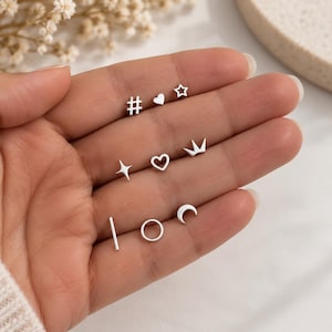 May include: A collection of nine small silver stud earrings arranged in three rows. The shapes include a hashtag, a solid heart, a star, a four-pointed star, an open heart, a crown, a vertical bar, a circle, and a crescent moon symbol.