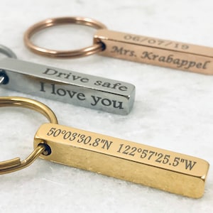 May include: Three gold, silver and rose gold bar keyrings with engraved messages. The gold keyring has the coordinates 50°03'30.8"N 122°57'25.5"W engraved on it. The silver keyring has the message "Drive safe I love you" engraved on it. The rose gold keyring has the date 06/07/19 and the name "Mrs. Krabappel" engraved on it.