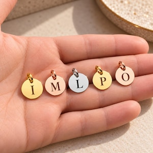 May include: Five small circular metal charms, each featuring a single engraved letter. The charms are arranged in a row and include gold, rose gold, and silver tones, with the letters I, M, L, P, and O clearly visible on their surfaces.