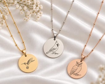Könnte beinhalten: Drei runde Anhänger-Halsketten an zarten Erbsketten mit Kügelchen. Der goldene Anhänger zeigt einen Kolibri, der silberne einen Kardinal und der roségoldene eine Eule. Jeder runde Anhänger ist mit einem detaillierten Vogel graviert.