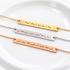 May include: Three horizontal bar necklaces in gold, silver, and rose gold tones. Each metal bar features engraved Morse code dots and dashes. The necklaces are attached to delicate matching metal chains, displaying a clean and polished finish.