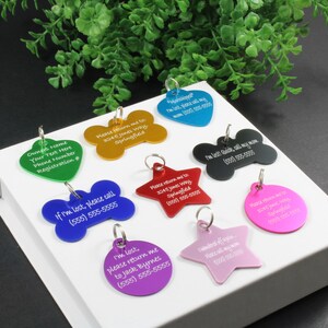 May include: A collection of nine colourful metal pet ID tags in various shapes including bones, stars, hearts, and circles. Each tag features engraved white text with contact information and phrases like 'If I'm lost, please call' and 'Please return me to'.