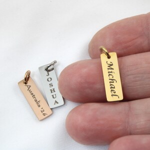 May include: Three rectangular metal charms with engraved text. One gold charm reads "Michael", a silver charm reads "Joshua", and a rose gold charm reads "Australia '26". Each charm has a small loop for attachment.