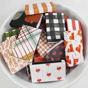 May include: A bowl filled with matchboxes featuring various autumnal designs. The matchboxes have patterns of plaid, gingham, leaves, pumpkins, and hearts. The matchboxes are in shades of orange, red, green, black, and white.