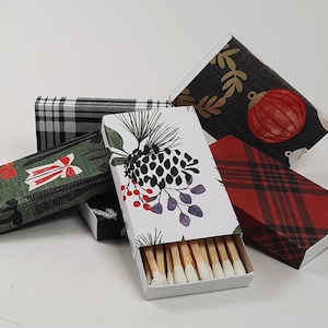 May include: A collection of decorative matchboxes with various holiday-themed designs. The matchboxes feature patterns like plaid, pinecones, bows, and ornaments. The matches inside have white tips.