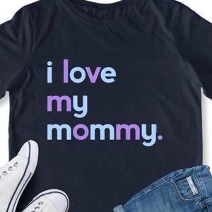 May include: Navy blue t-shirt with the text "i love my mommy" in light blue and purple. The shirt is paired with white sneakers and blue jeans.