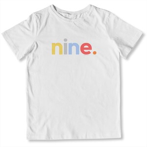 May include: White t-shirt with the word "nine." printed across the front in yellow, blue, and red. The shirt has short sleeves and a crew neck. The text is in a playful, colorful font.
