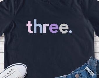 May include: Navy blue t-shirt with the word "three" in pastel colors. The shirt is paired with white sneakers and denim shorts, suggesting a child's outfit. A simple, yet stylish design for a birthday or special occasion.