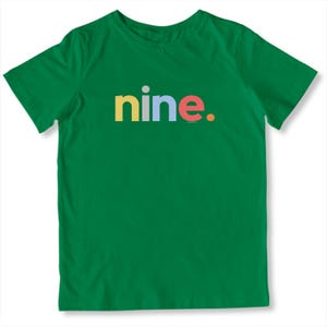 May include: A green t-shirt with the word "nine" in colorful letters. The word is in yellow, blue, and red. A small gray dot is above the "i" in "nine". The shirt is made of a soft material.