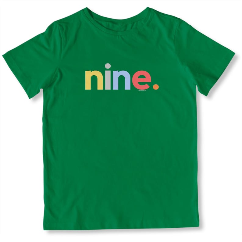 May include: A green t-shirt with the word "nine" in colorful letters. The word is in yellow, blue, and red. A small gray dot is above the "i" in "nine". The shirt is made of a soft material.