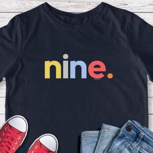 May include: Navy blue t-shirt with the word "nine" in colorful block letters. The word is in yellow, gray, blue, and red, with a small orange dot at the end. The shirt is paired with red sneakers and blue jeans.