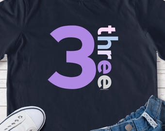 May include: A dark navy blue t-shirt featuring a large lavender number three on the left and the word three written vertically in light pink, light blue, and gray letters on the right, accompanied by white sneakers and blue denim jeans.