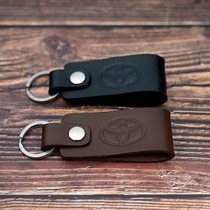 May include: Two leather keyrings with a silver key ring. One keyring is brown leather with a silver button and the other is black leather with a silver button. Both keyrings have a silver embossed Toyota logo.