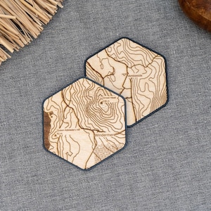 May include: Two hexagonal wooden coasters with a topographic map design. The coasters are light brown with dark brown outlines and intricate line details. The coasters are placed on a gray fabric surface, with decorative elements in the background.