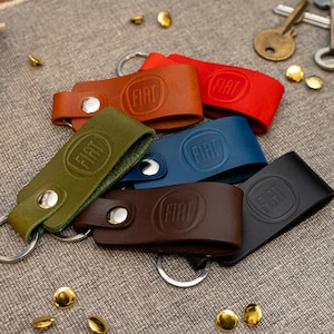 May include: A selection of leather keyrings in shades of green, brown, red, and blue. Each keyring has a silver ring and a debossed "FIAT" logo. The keyrings are displayed on a grey fabric surface, with scattered gold-coloured studs.