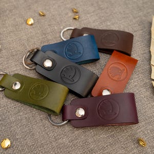 May include: A collection of six leather keychains in olive green, black, blue, dark brown, tan, and burgundy. Each keychain features a silver metal split ring, a silver snap button, and a circular embossed logo of a winged arrow.