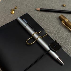 May include: A black notebook featuring a silver pen held in place by a gold-toned metal clip and a black elastic band. The notebook has a smooth, matte cover, and the pen is secured horizontally across the front, providing a functional and organised stationery setup for writing and note-taking tasks.