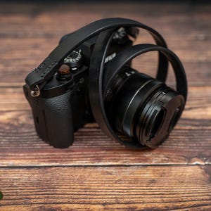 May include: A black camera with a black leather strap. The camera is sitting on a wooden surface.