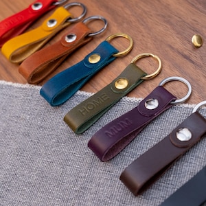 May include: A selection of leather keyrings in a range of colours, including red, yellow, brown, blue, green, purple, and dark brown. Some keyrings have embossed text, such as "HOME" and "MUM". Each keyring has a metal ring for keys.