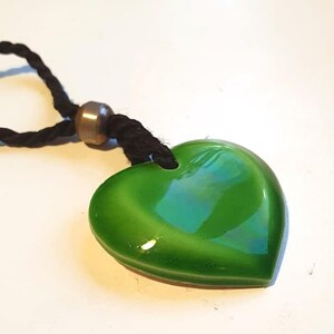 Könnte beinhalten: Ein grüner herzförmiger Anhänger mit glänzendem Finish, der an einer schwarzen Kordelkette hängt. Der Anhänger hat eine glatte, abgerundete Form und eine metallische Perle. Die Halskette ist ein schlichtes, elegantes Accessoire.