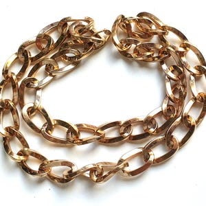 Könnte beinhalten: Goldfarbene Kette mit großen ovalen Gliedern. Die Halskette besteht aus Metall und hat ein glänzendes Finish. Die Kette ist in einer gebogenen Form angeordnet, geeignet zum Tragen um den Hals. Ein auffälliges Schmuckstück.