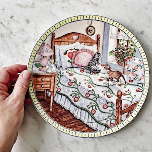 May include: A ceramic plate featuring a colourful illustration of a bedroom scene with a bed, two cats, a bedside table and a lamp. The bed has a white and blue quilt with red flowers. The cats are sitting on the bed and the bedside table has a lamp with a pink shade.